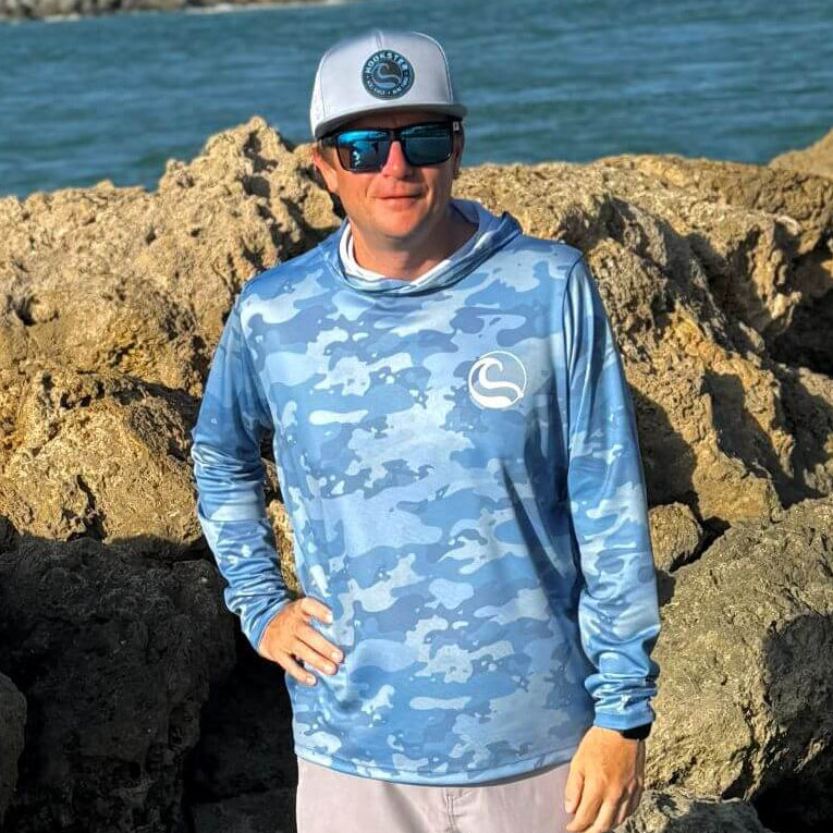 Person standing on rocks by the ocean wearing a blue camouflage shirt and white cap.