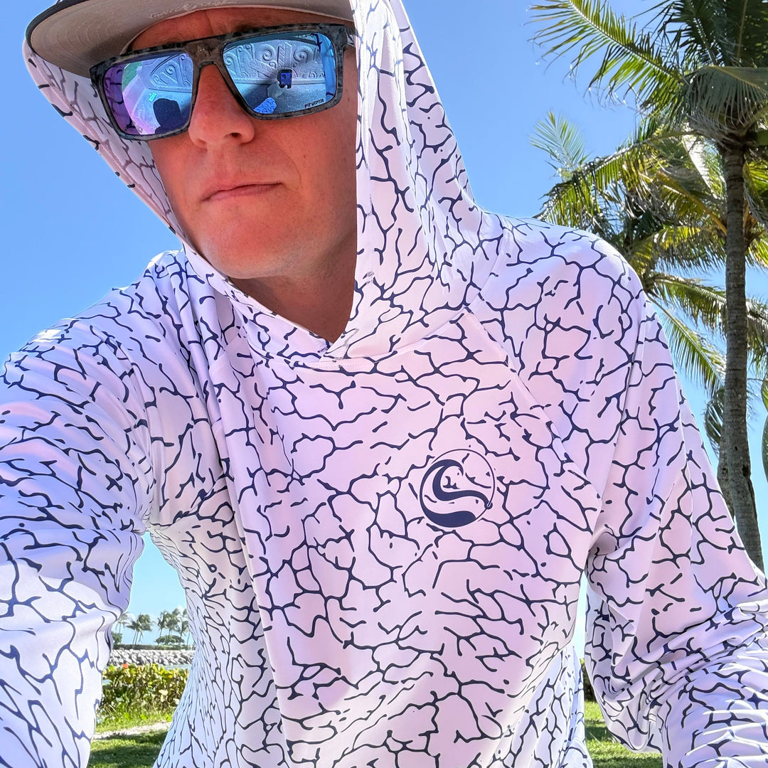 Angler in sunglasses taking a selfie under palm trees, wearing the HOOKSTER Tide Trace UPF 50+ UV hoodie with the hood up, white with a navy cracked-earth print