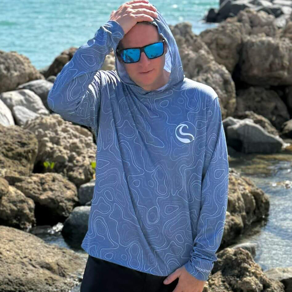 Man wearing the Topo Slate Men's UV Hoodie with hood up and sunglasses, standing on rocks by the water