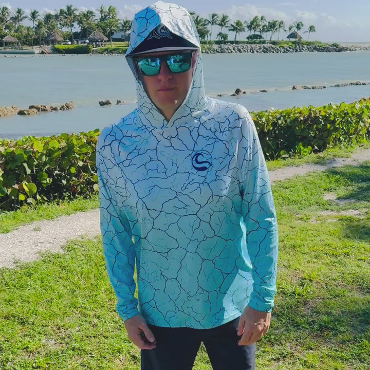 Video of an angler on the waterfront turning to show the front, back and thumbhole cuffs of the HOOKSTER Reef Fade UPF 50+ UV hoodie, white fading to aqua with a navy cracked-earth print
